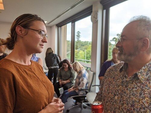 A woman and an older man talking in a cream-walled room, near a window overlooking a park