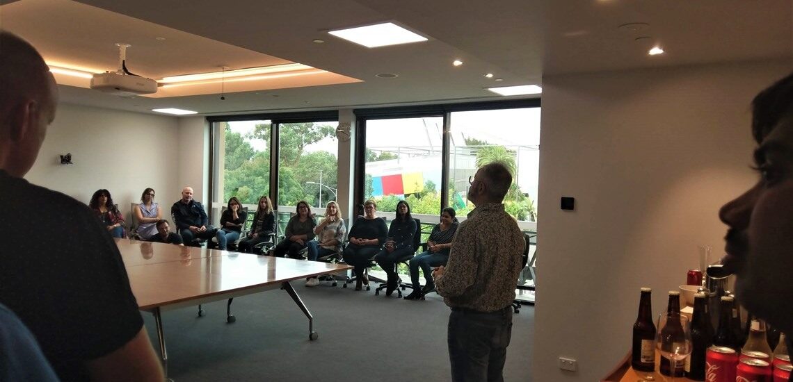 A older man presenting to a group of people in a cream-walled room overlooking a park.